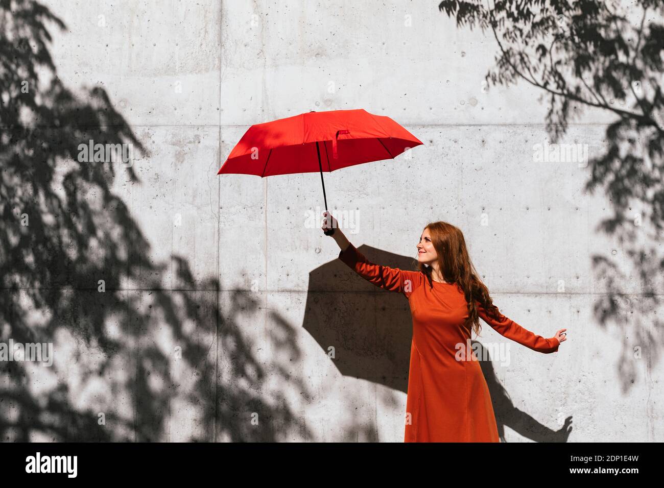 Smiling redhead woman holding umbrella while standing against tree shadow wall Foto Stock