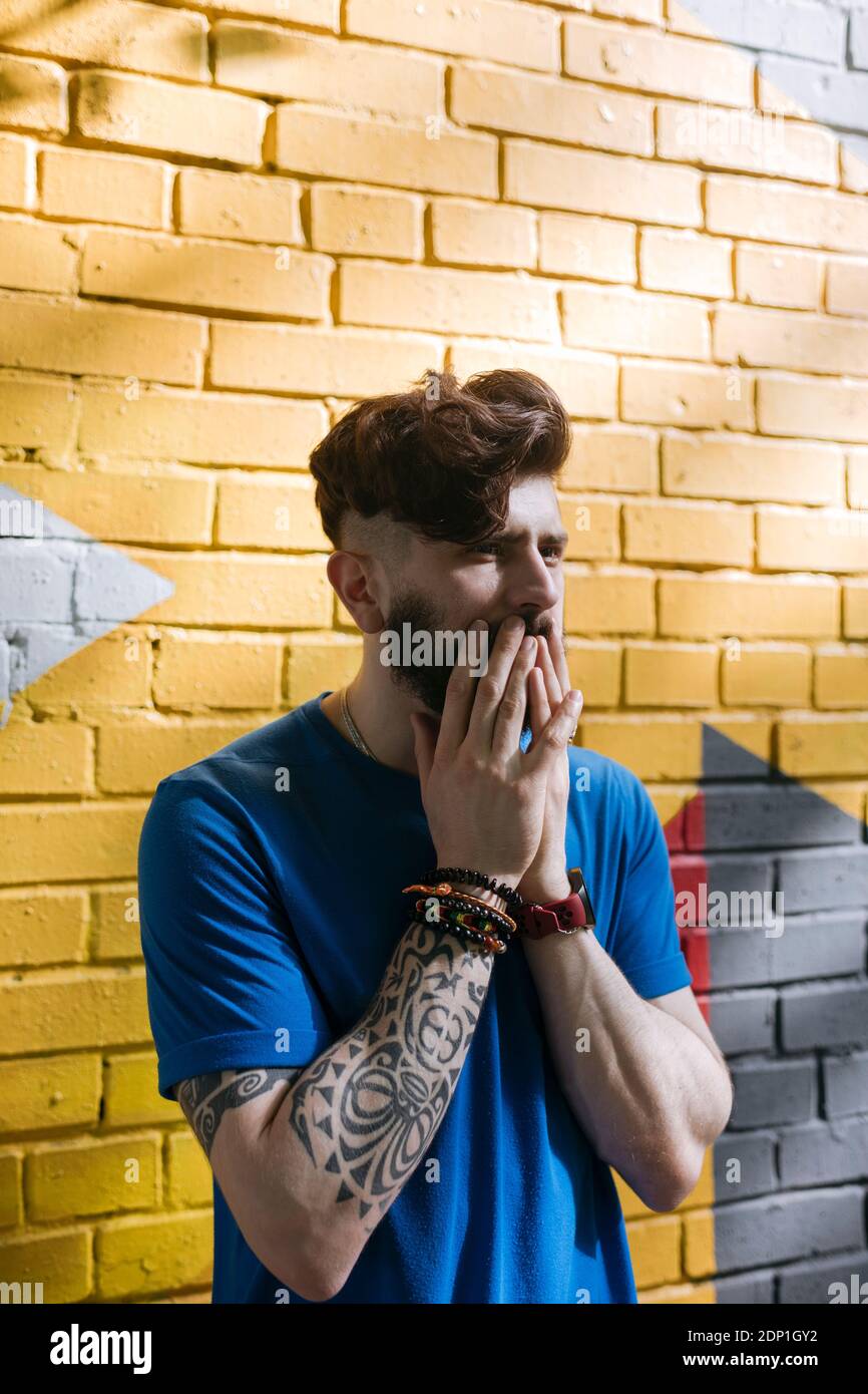 Giovane uomo con tatuaggio in piedi davanti al muro di mattoni gialli, coprendo la bocca con le mani Foto Stock