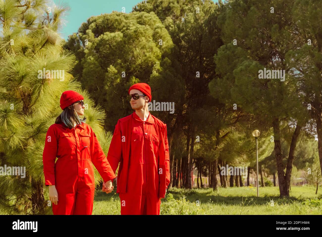 Giovane coppia che indossa tute rosse e cappelli in piedi mano mano su un prato che si guarda l'un l'altro Foto Stock