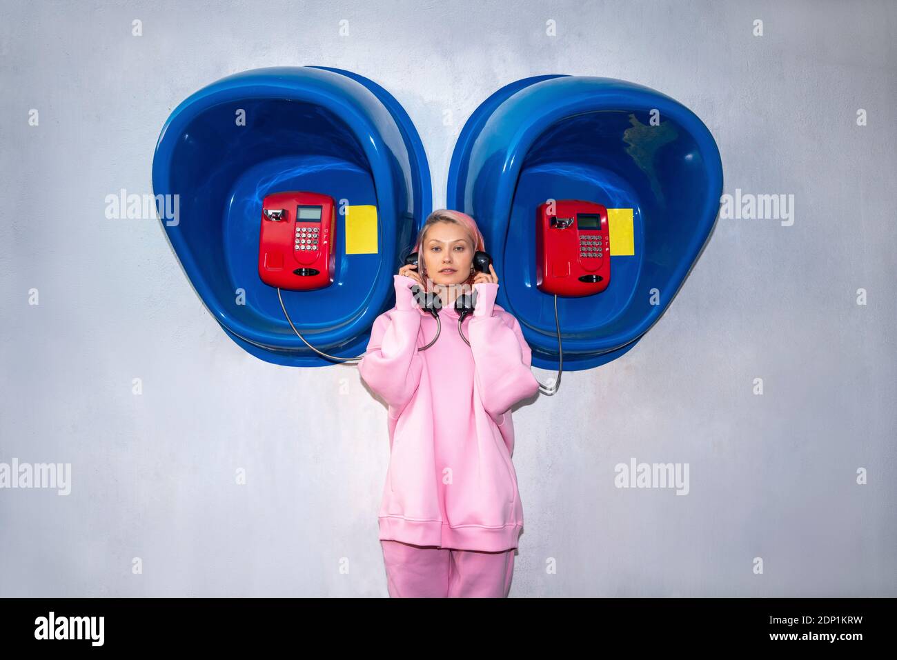 Giovane donna con capelli rosa che indossa una camicia con cappuccio rosa in piedi davanti alle cabine telefoniche e ai ricevitori Foto Stock