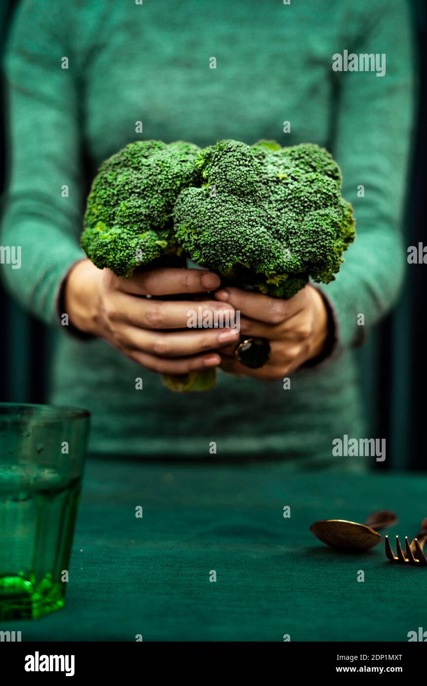 Sezione mediana della donna in piedi con broccoli e cucchiaio, forchetta, vetro tenuto sul tavolo Foto Stock
