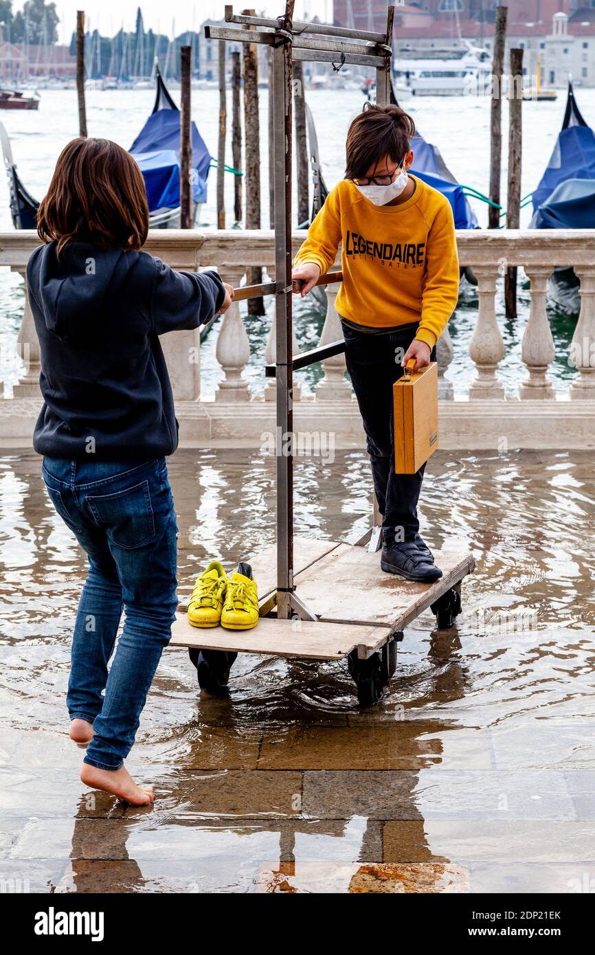 Due bambini giocano con un tram per bagagli dell'hotel durante l'acqua alta (alta marea) Piazza San Marco, Venezia, Italia. Foto Stock