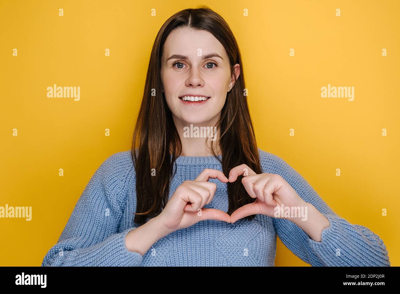 Allegra giovane donna carina con ampio sorriso affascinante mettere le mani sul petto fare con le dita forma del cuore simbolo di carità, isolato su studio giallo Foto Stock
