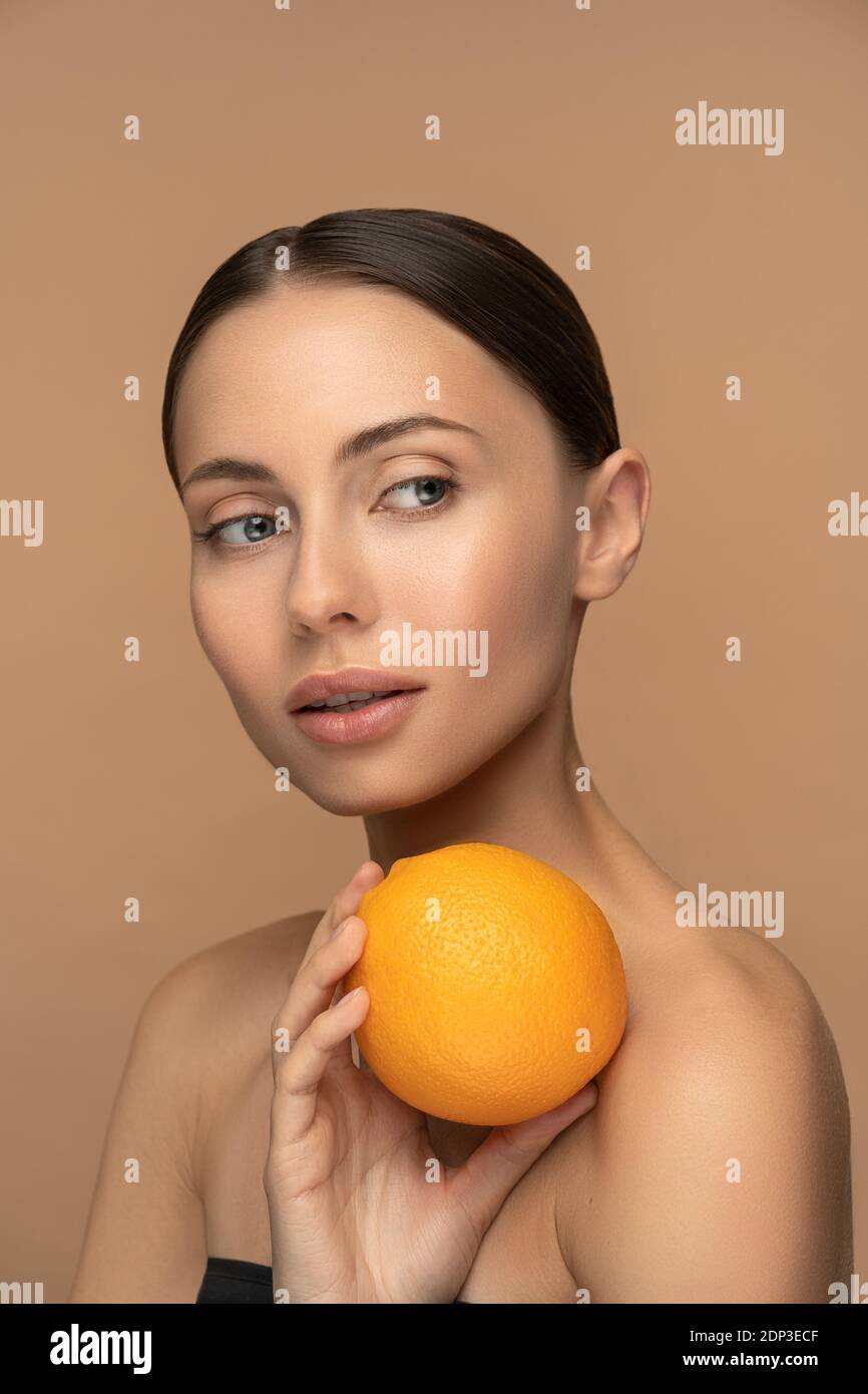 Donna con perfetta pelle del viso, capelli pettinati, che tiene arancione. Ritratto di donna con trucco naturale e agrumi in posa su sfondo beige. Vitam Foto Stock