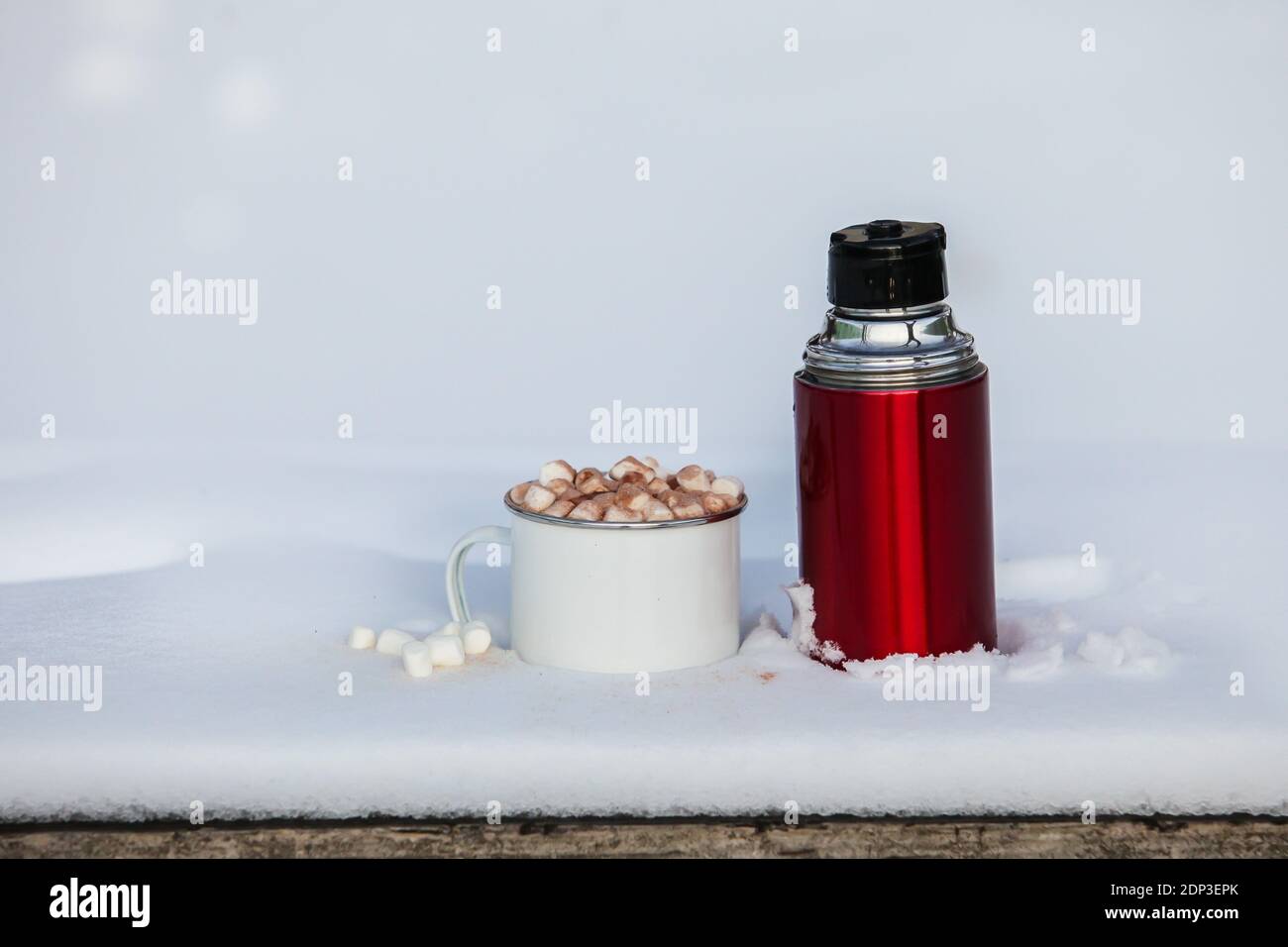 Thermos rossi prossima tazza con cioccolato caldo con marshmallows sopra tavolo con neve all'esterno Foto Stock