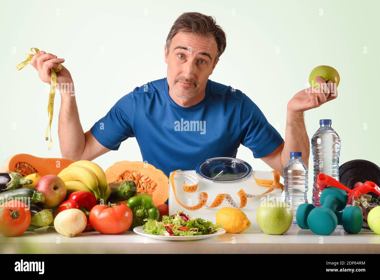 Uomo espressivo in abbigliamento sportivo pronto a mantenere una vita sana con cibo sano e accessori per lo sport su un tavolo e sfondo verde isolato Foto Stock