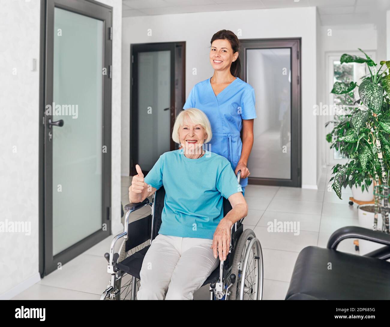 Cordiale infermiera geriatrica che assiste una donna anziana in sedia a rotelle nel corridoio della casa di cura. assistenza medica per le persone con disabilità Foto Stock
