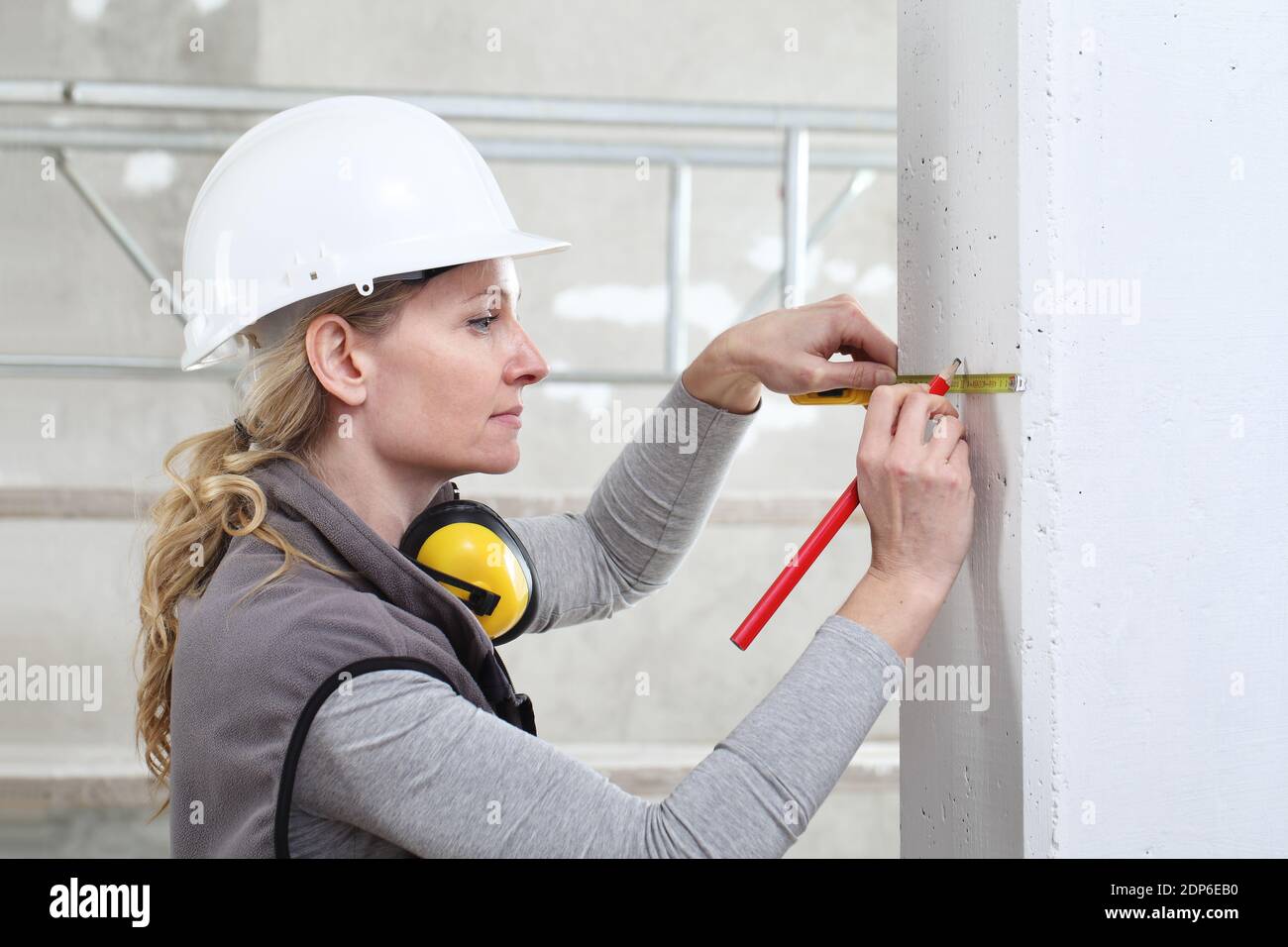 donna operatrice di costruzione lavora con metro a nastro e matita, misurare la parete in cantiere, indossare casco e cuffie di protezione dell'udito Foto Stock