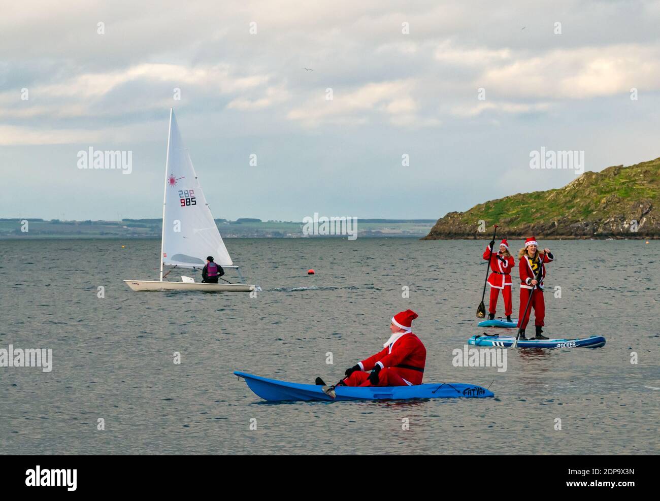 North Berwick, East Lothian, Scozia, Regno Unito, 19 dicembre 2020. Paddle imbarco Santas per la carità: Un'iniziativa locale della comunità da North Berwick News e opinioni chiamato 'Christmas Cheer' raccoglie oltre £5,000 fondi per le famiglie in bisogno. I paddle boarder sono vestiti in costumi di Santa con una barca a vela che passa accanto Foto Stock