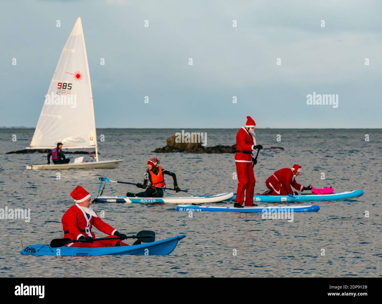 North Berwick, East Lothian, Scozia, Regno Unito, 19 dicembre 2020. Paddle imbarco Santas per la carità: Un'iniziativa locale della comunità da North Berwick News e opinioni chiamato 'Christmas Cheer' raccoglie oltre £5,000 fondi per le famiglie in bisogno. I paddle boarder sono vestiti in costumi di Santa con una barca a vela che passa accanto Foto Stock