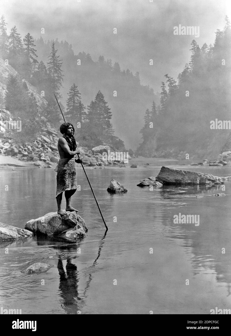 Iconica fotografia d'epoca di Edward Curtis intitolata A Smoky Day at the Sugar Bowl UN uomo di Hupa si erge su una roccia in un lago con uno sfondo di foresta. Foto Stock