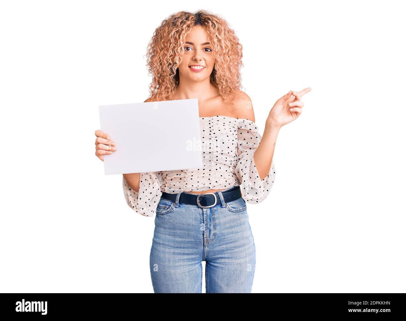 Giovane donna bionda con capelli ricci che tengono in vendita poster sorridente felice di puntare con mano e dito a lato Foto Stock