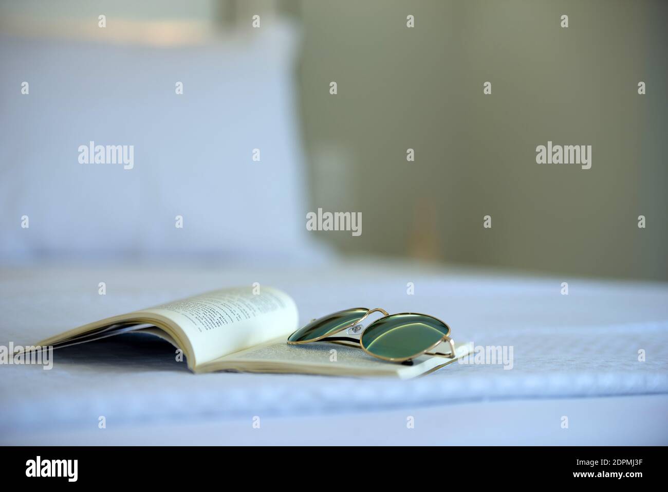 Closeup di libro aperto e occhiali da sole che si posa su bianco letto sullo sfondo sfocato della camera da letto Foto Stock