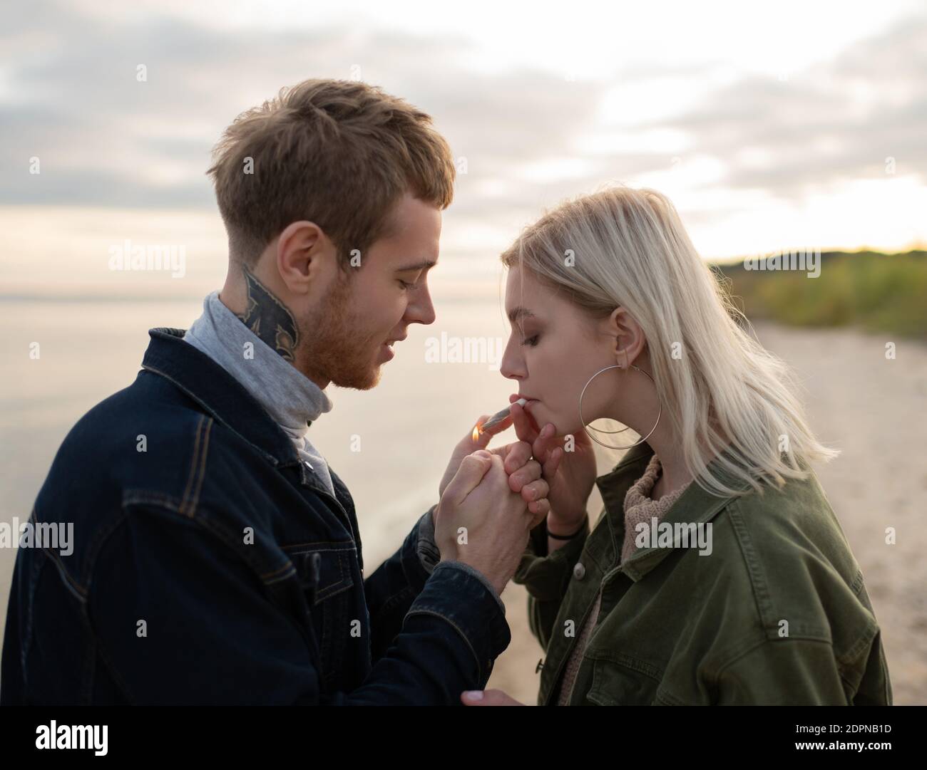 Vista laterale dell'uomo tatuato che illumina la sigaretta di cannabis mentre è biondo donna che inala fumo mentre si rilassa nella natura autunnale in serata Foto Stock Vista laterale dell'uomo tatuato che illumina la sigaretta di cannabis mentre è biondo donna che inala fumo mentre si rilassa nella natura autunnale in serata Foto Stock