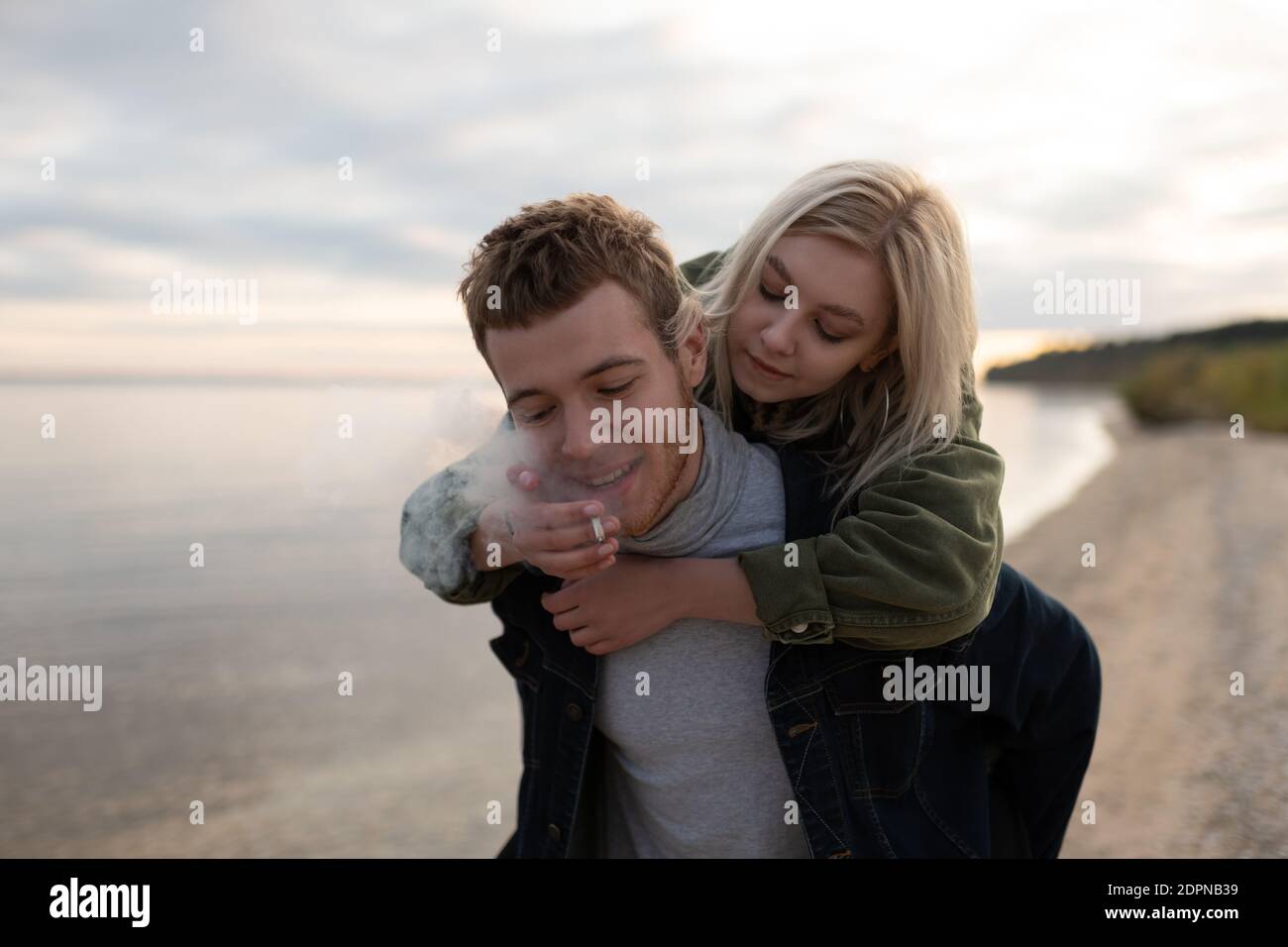 Un ragazzo hipster sorridente che dà un giro in piggyback a una ragazza che condivide la cannabis smussate mentre camminate da soli sulla costa autunnale Foto Stock