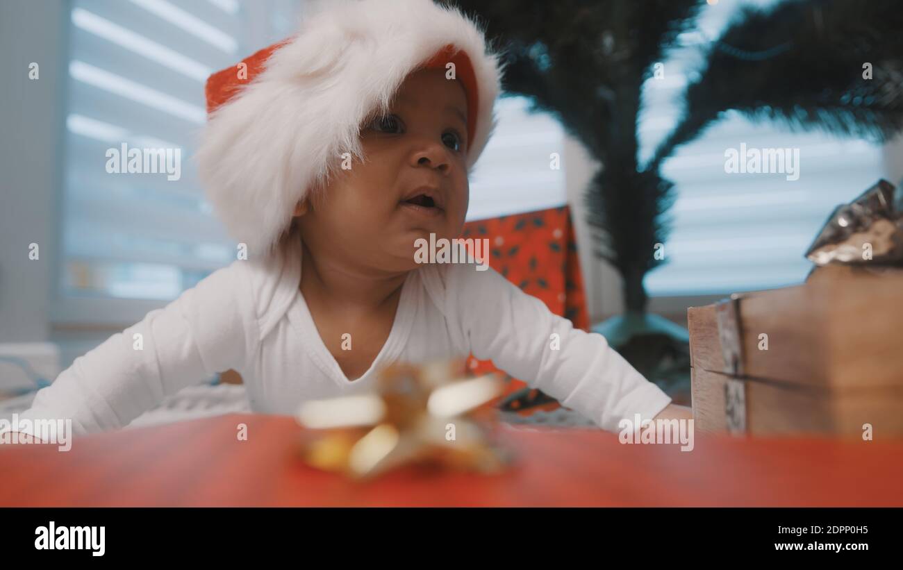 Cute bambino di pelle scura con il cappello di santa circondato dai regali che hanno il tempo della pancia sotto l'albero di natale. Foto di alta qualità Foto Stock