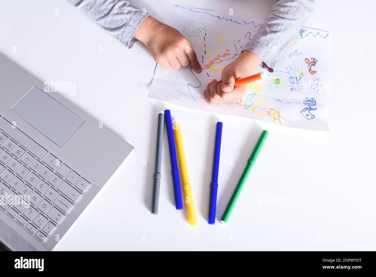 vista orizzontale dall'alto delle mani di un ragazzo caucasico che disegnano su un foglio con penne in feltro su un tavolo bianco con un computer portatile. Concetto di scuola domestica Foto Stock