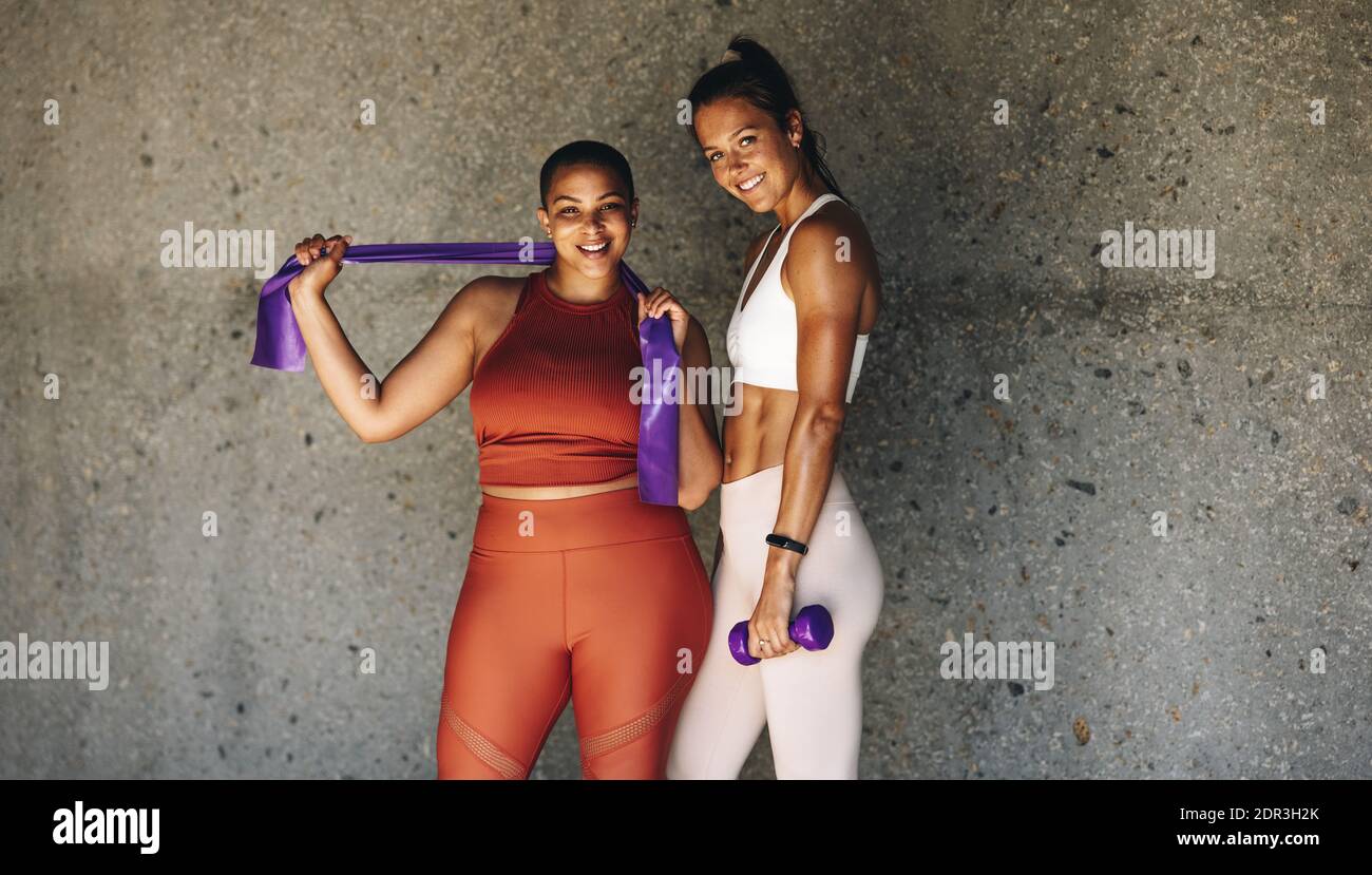 Due donne che si esercitano insieme con la banda di resistenza e dumbbell. Amici femminili in sportswear reggendo la banda di resistenza e dumbbell guardando la macchina fotografica Foto Stock