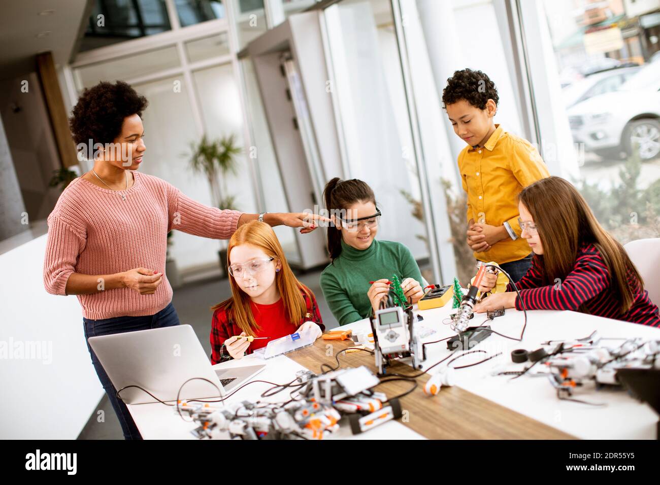 Gruppo di bambini felici con la loro scienza femminile afroamericana insegnante con i giocattoli elettrici di programmazione del laptop e robot alla robotica in aula Foto Stock