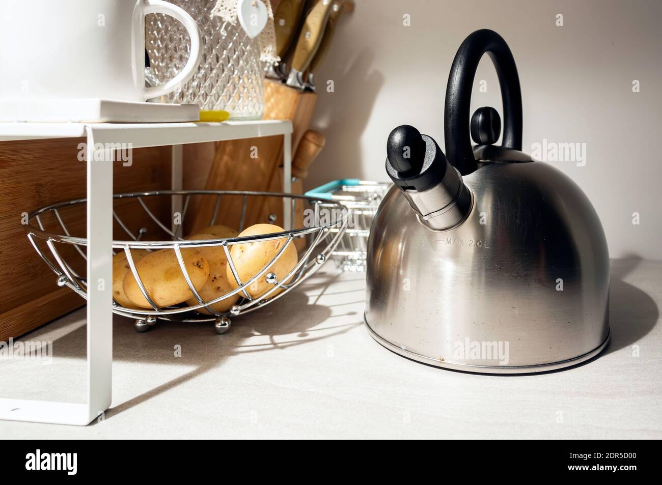 Moderno bollitore d'acciaio accanto ad un cesto di patate in una cucina domestica scarsamente illuminata. Accessori per la vita domestica e la cucina Foto Stock