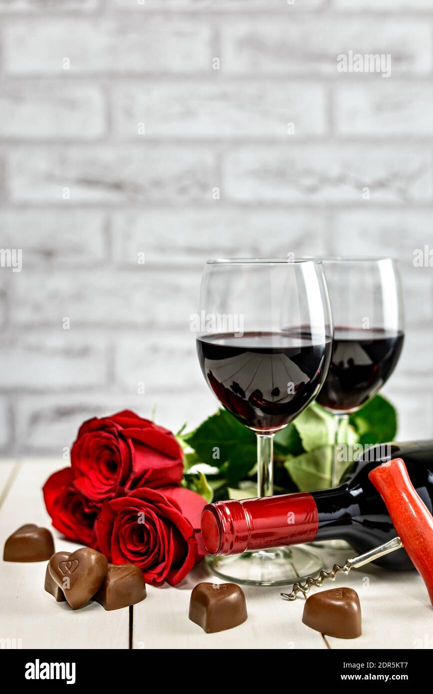 Concetto di San Valentino. Due bicchieri di vino, bottiglia di vino, cavatappi, rose rosse e cuori di cioccolato su un tavolo di legno bianco con spazio copia per il testo. Foto Stock