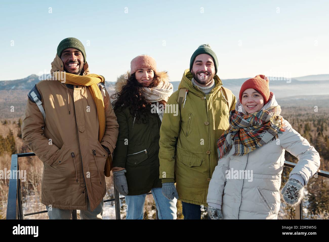 Felici giovani amici interculturali in abbigliamento da inverno in piedi di fronte fotocamera contro il cielo blu sulle montagne coperte di neve mentre raffreddamento fuori Foto Stock