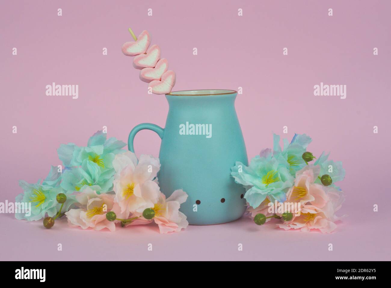 Una tazza di ceramica blu con cioccolato caldo e marshmallow su un bastone, con fiori su sfondo rosa Foto Stock