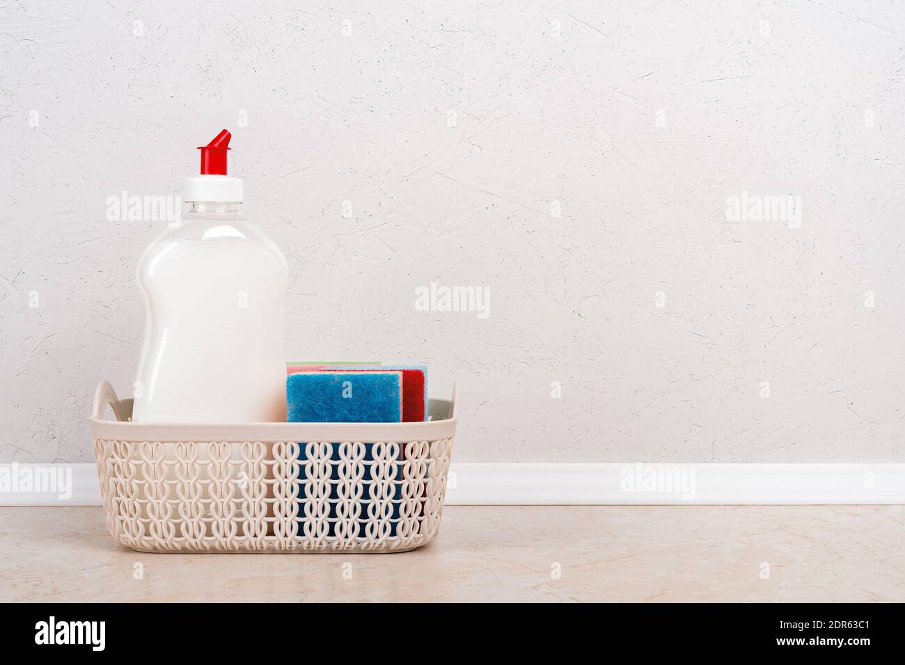 Liquido bianco per il lavaggio delle stoviglie e alcune spugne in schiuma in un piccolo cestello sul tavolo da cucina contro la parete bianca in cemento. Detergente per uso domestico. Spazio di copia. Foto Stock