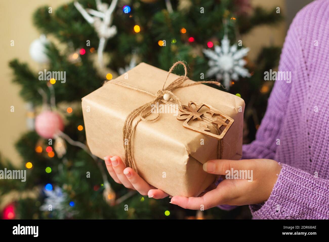 Le mani di donne che tengono il presente piacevole avvolto in carta artigianale, albero di Natale sullo sfondo, concetto di spreco zero Foto Stock