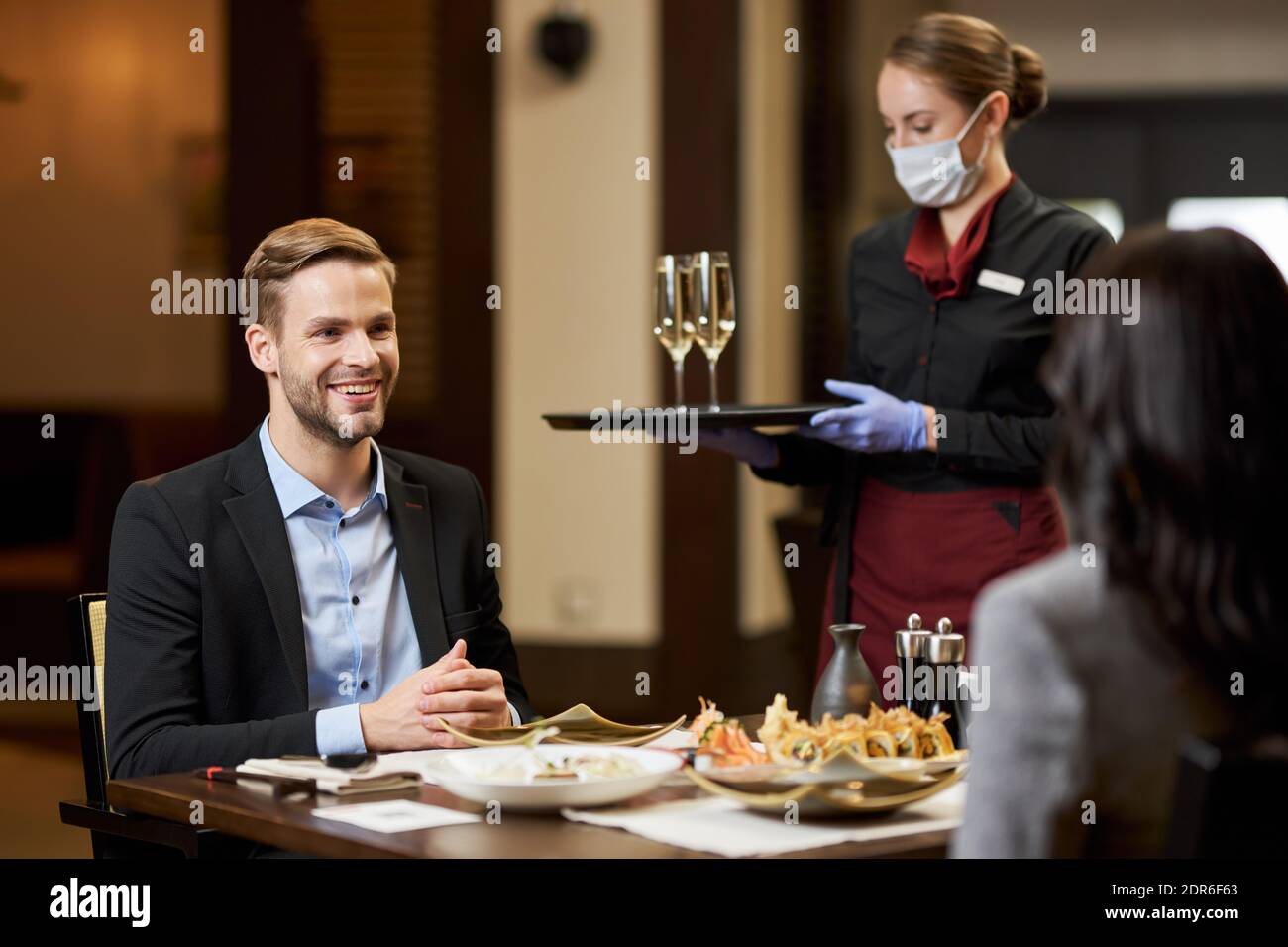Giovane attraente che ha una conversazione con una donna attraverso tavolo mentre cameriera in maschera portando ordine pasto Foto Stock