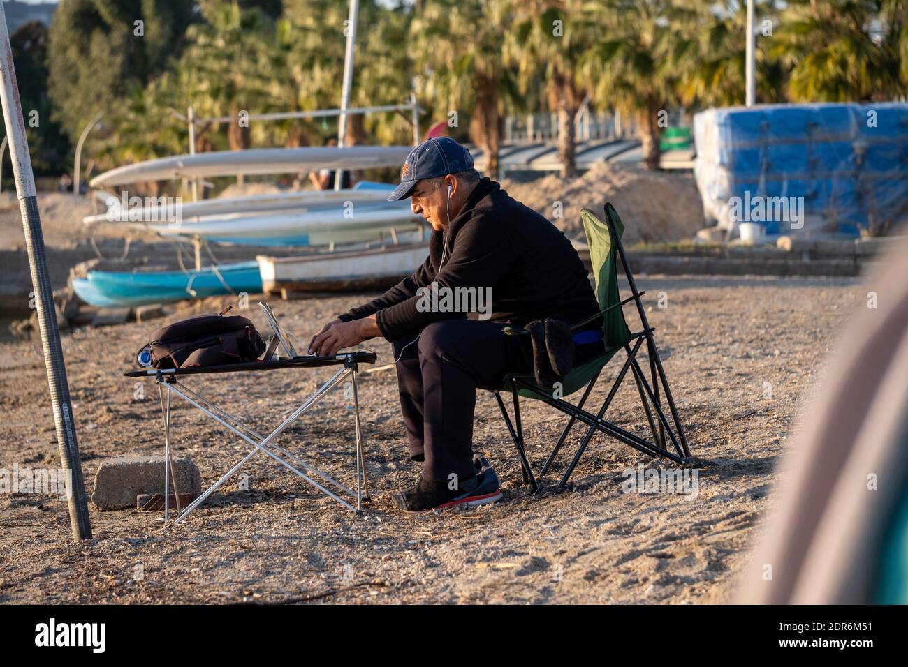 Mugla, Turchia, 12.03.2020: Un uomo con cuffia si siede sulla riva e lavora sul suo laptop che si trova su un tavolo da campeggio. In palme di sfondo. Foto Stock
