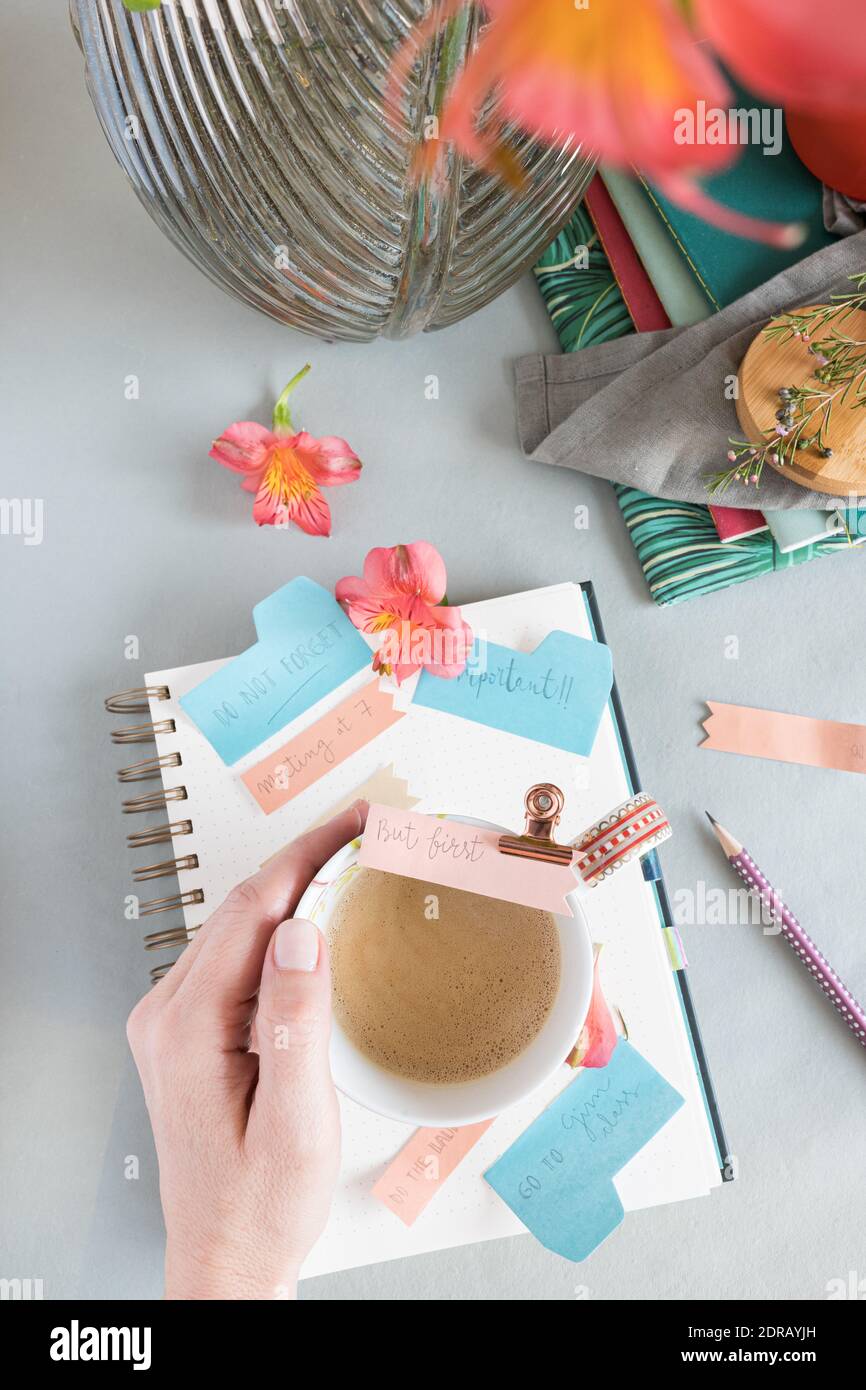 Mano donna che tiene un caffè schiumoso su un notebook a spirale pieno di note di promemoria multicolore su uno sfondo pulito. Foto Stock