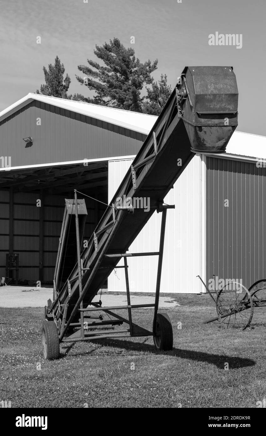 Primo piano vista astratta di un attrezzo agricolo con elevatore di fieno e grano stagionato, con vista di un grande e moderno capannone macchina e sfondo blu cielo Foto Stock
