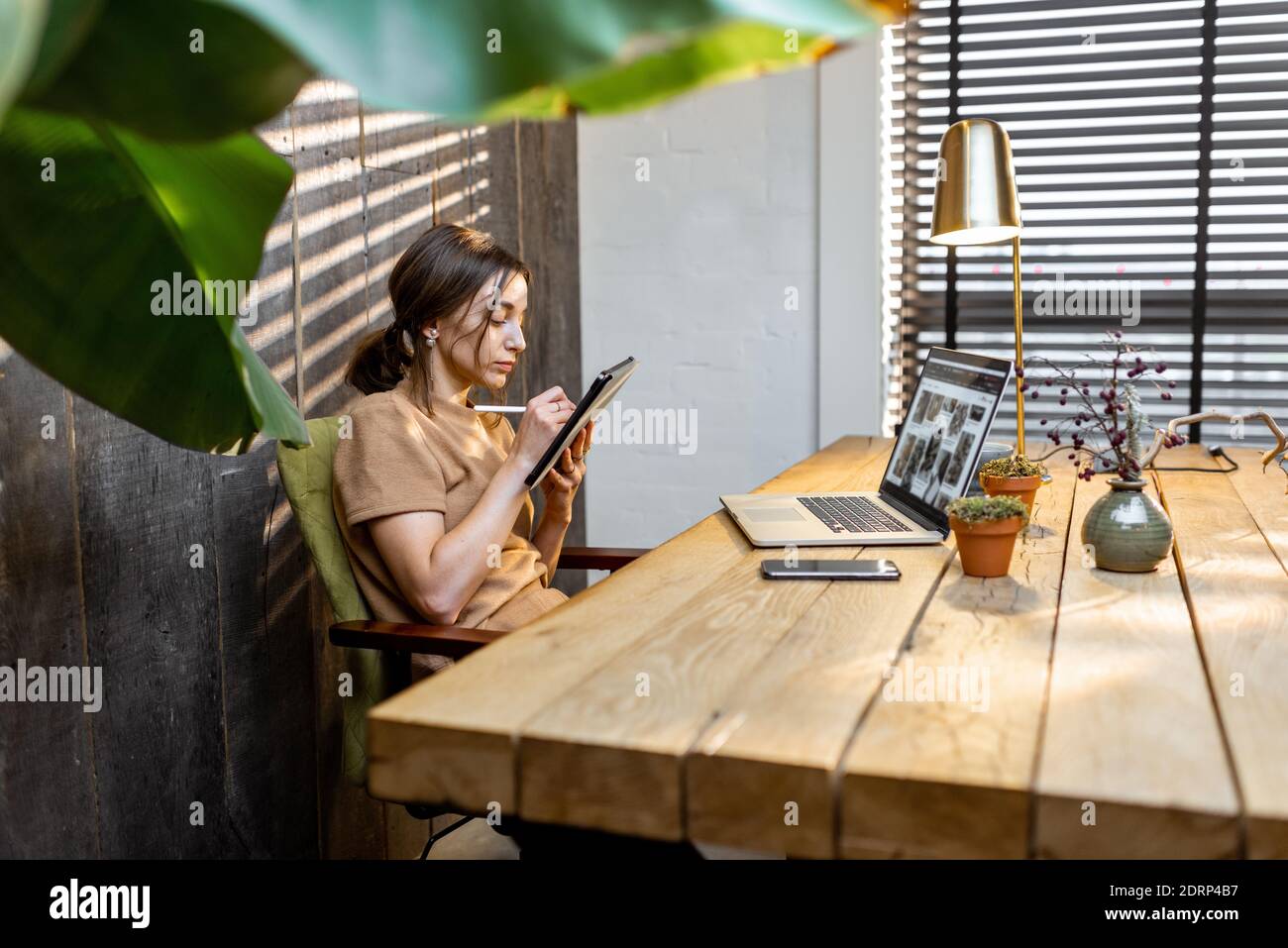 Donna creativa in comoda tuta domestica che lavora su tablet digitale nell'accogliente e elegante ufficio a casa Foto Stock