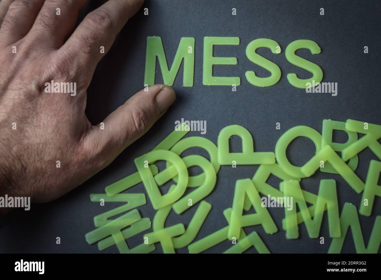 Mano umana accanto alla parola Mess scritto con lettere di plastica su sfondo grigio carta, con un mazzo di lettere in disordine, concetto Foto Stock