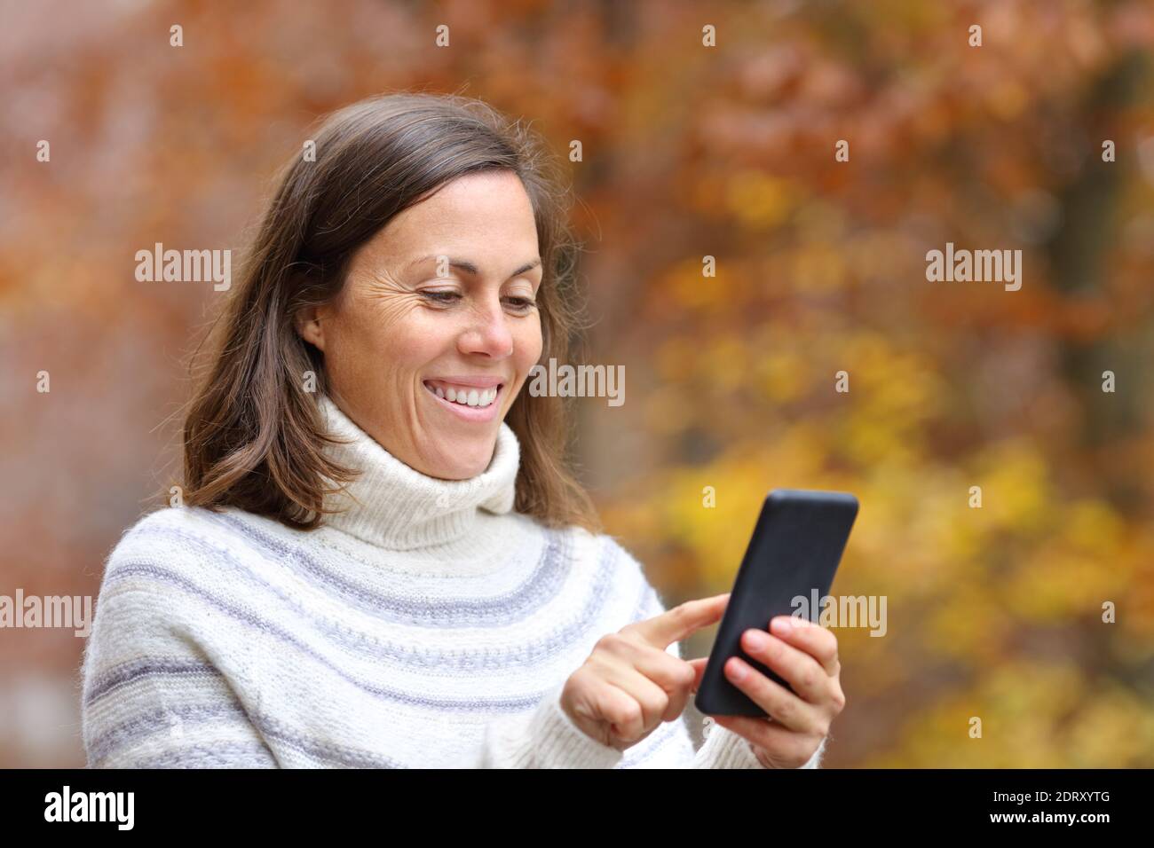 Donna adulta felice che usa lo smartphone camminando nella stagione autunnale in un parco Foto Stock