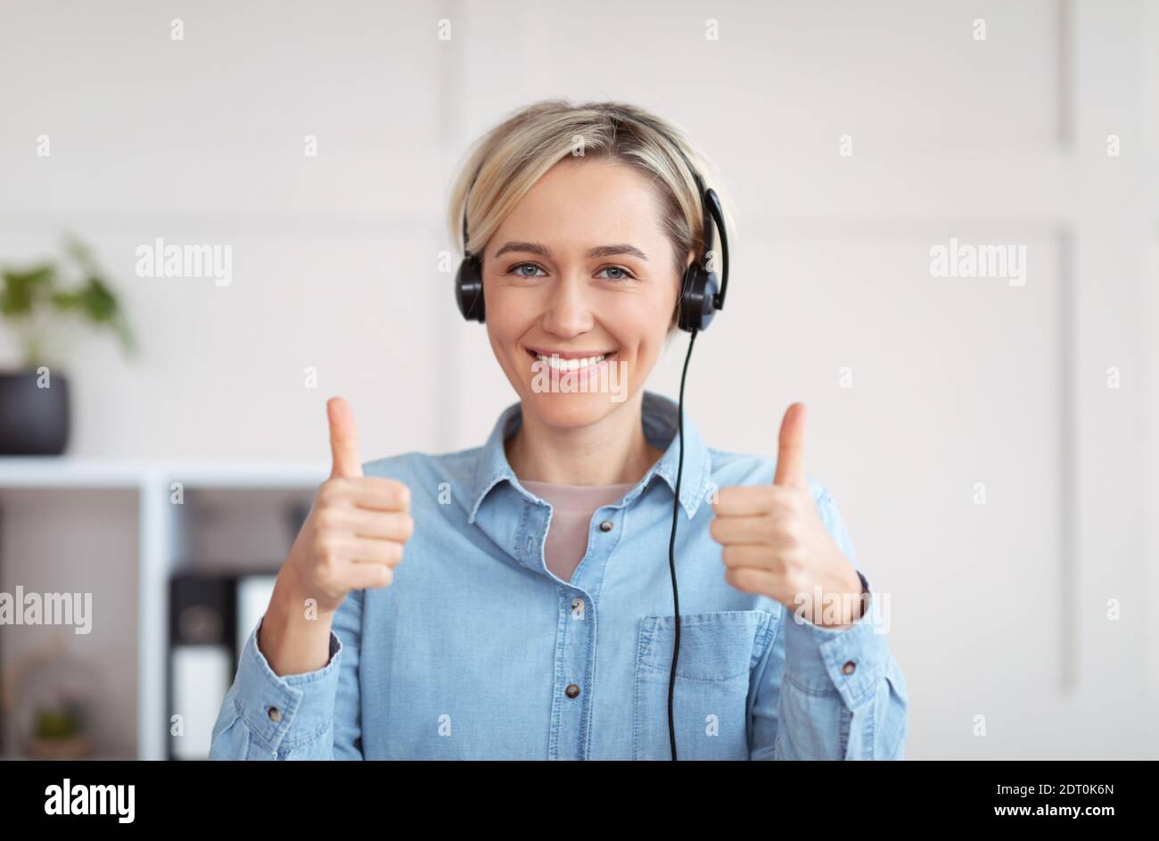 Istruzione, lavoro, business a distanza. Felice giovane donna in cuffia guardando la fotocamera e mostrando i pollici verso l'alto gesto Foto Stock