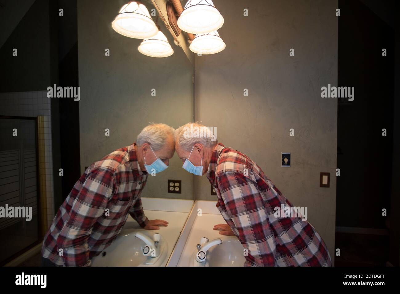 Uomo anziano in maschera protettiva Covid appoggiando la testa sullo specchio in bagno Foto Stock