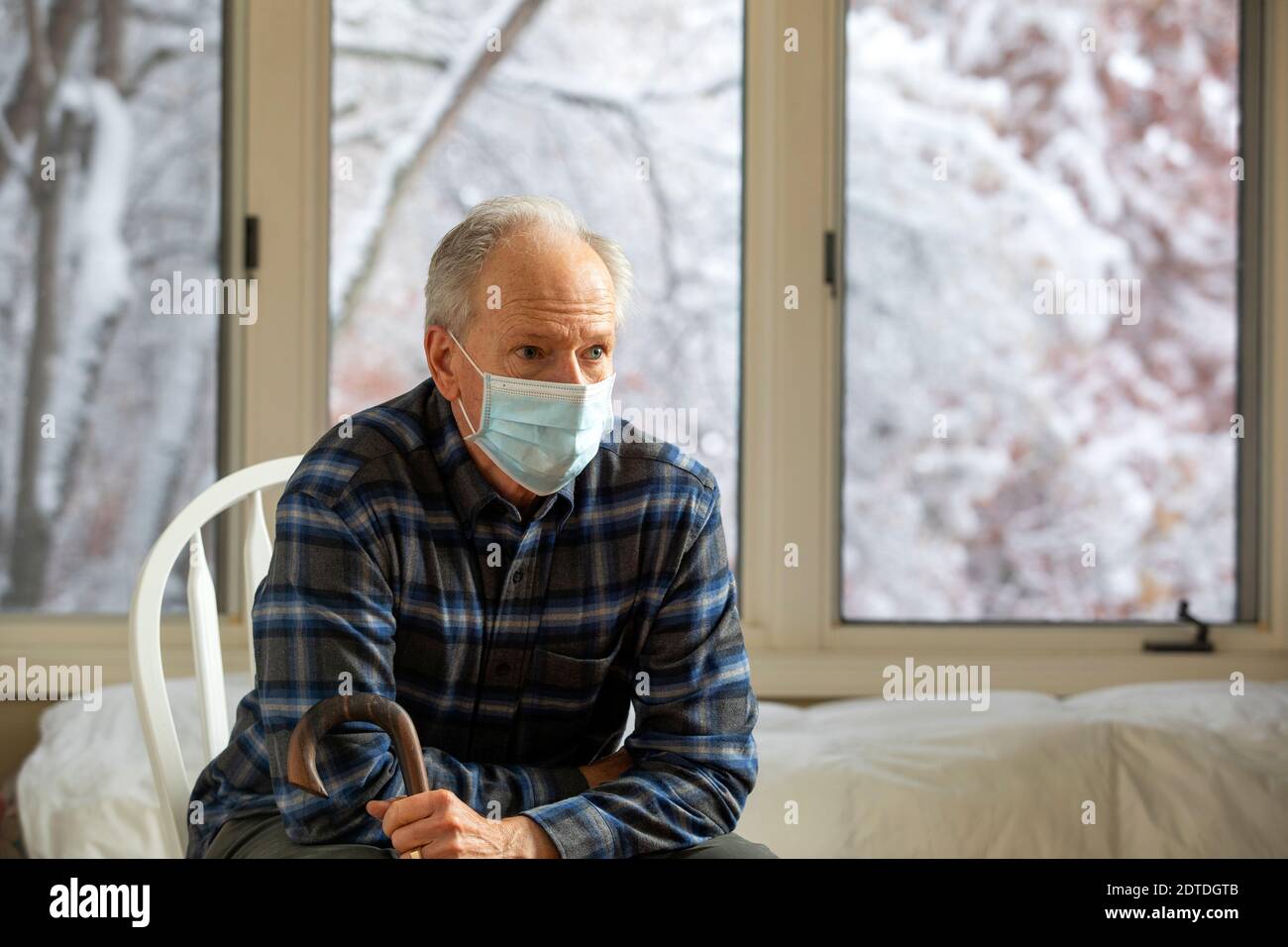 Uomo anziano in maschera di protezione Covid seduto sulla sedia vicino finestra e cane di tenuta Foto Stock