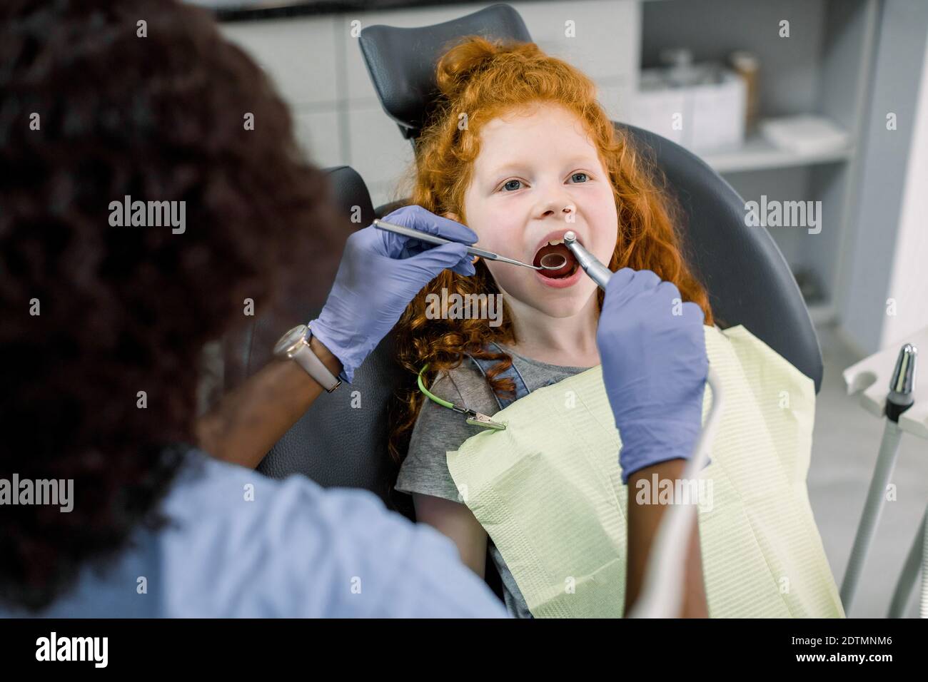 Carino piccola ragazza con capelli rossi graziosi durante la procedura dentale presso la clinica dentale dei bambini moderni. Dentista femminile per bambini donna africana che cura Foto Stock