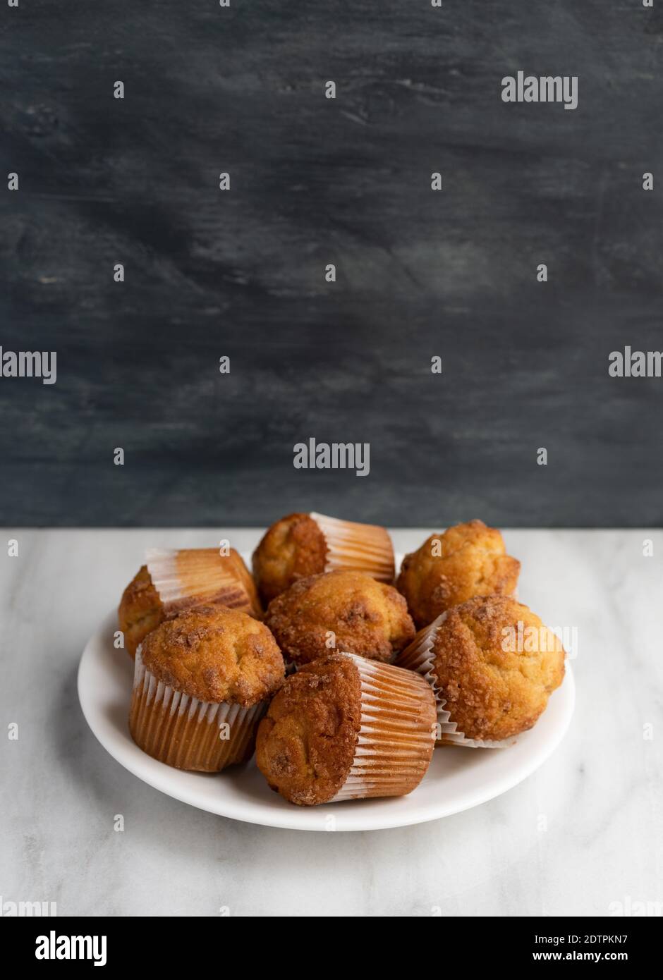 Vista laterale di diversi muffin Strausel a banana su una piastra bianca sopra un piano in marmo con una parete grigia sullo sfondo. Foto Stock