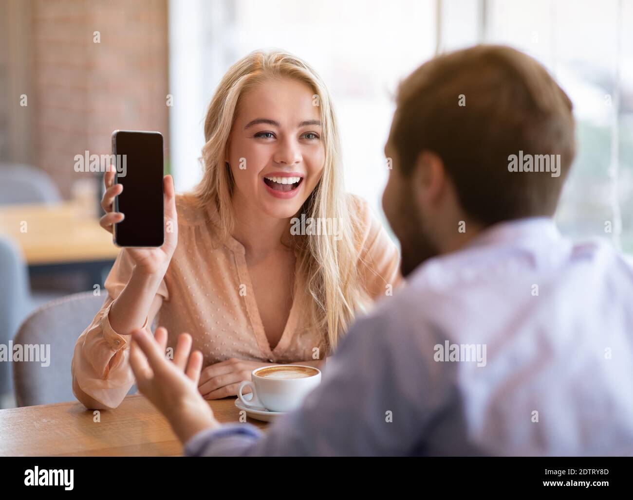 Giovane donna allegra che mostra il suo smartphone boyfriend con schermo vuoto al bar, mockup per il design Foto Stock