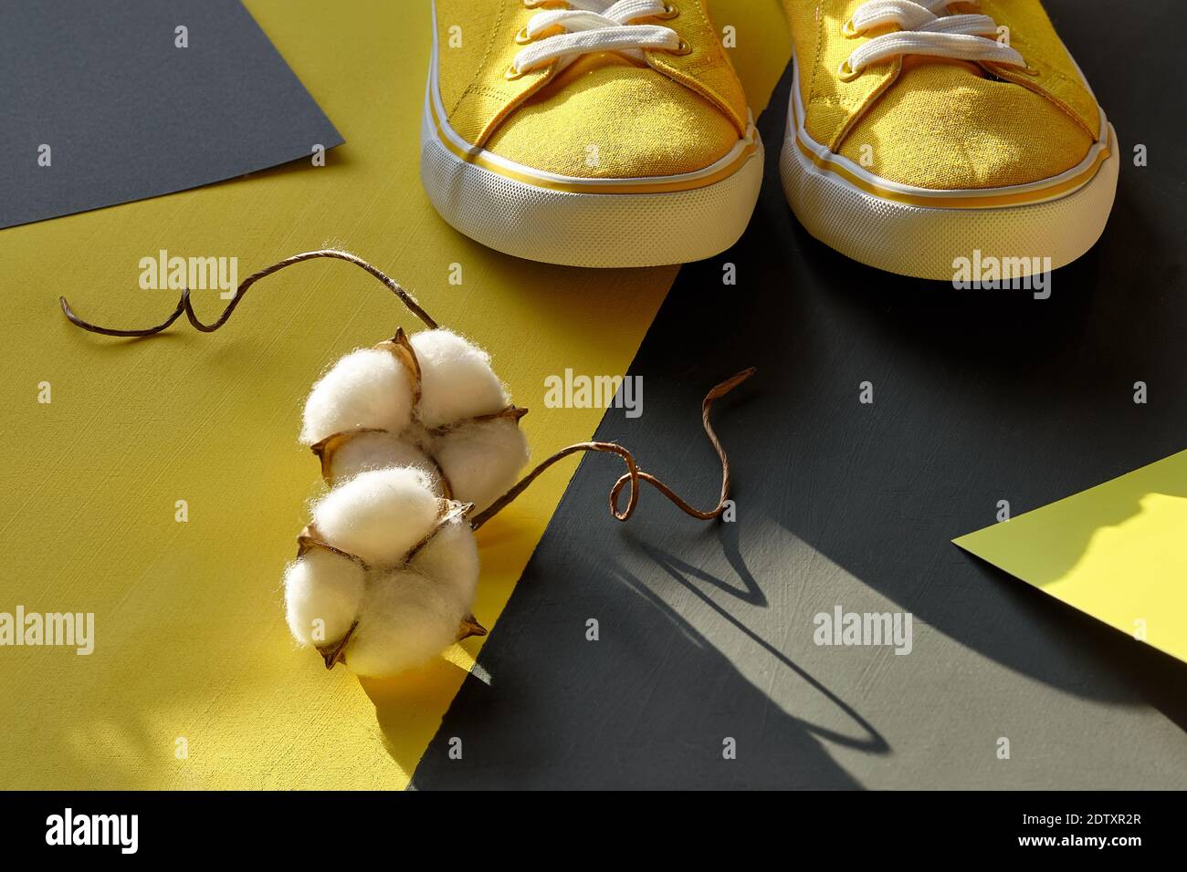 Paio di sneaker gialle e fiori decorativi in cotone su sfondo giallo grigio a strati. Giallo e grigio, colori di 2021. Foto Stock