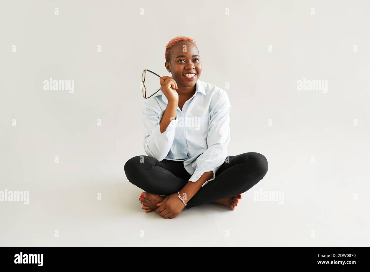 La giovane donna nera con i corti capelli rosa seduti il pavimento mantenendo i suoi occhiali e sorridendo Foto Stock