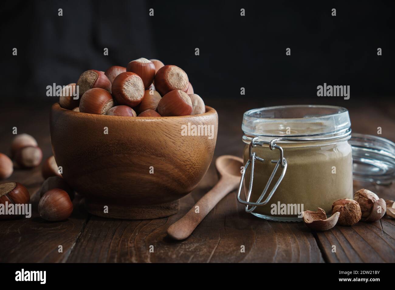 Ciotola in legno di nocciole, barattolo in vetro di burro di nocciole organico grezzo o pasta su tavola da cucina. Foto Stock