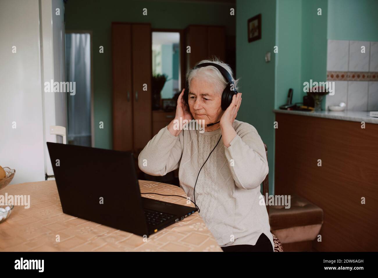 Una donna anziana con le cuffie seduto a un tavolo e che parla con la sua famiglia durante una videochiamata su un computer portatile. Isolamento della comunicazione durante COVID-19 Foto Stock