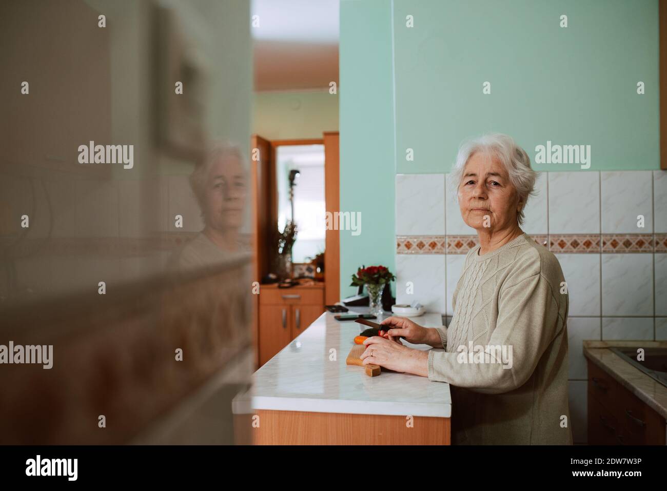 Carino nonna cucina pranzo e tritare le verdure sul tavolo da cucina. Una donna in pensione con capelli grigi mentre si cucina il pranzo Foto Stock
