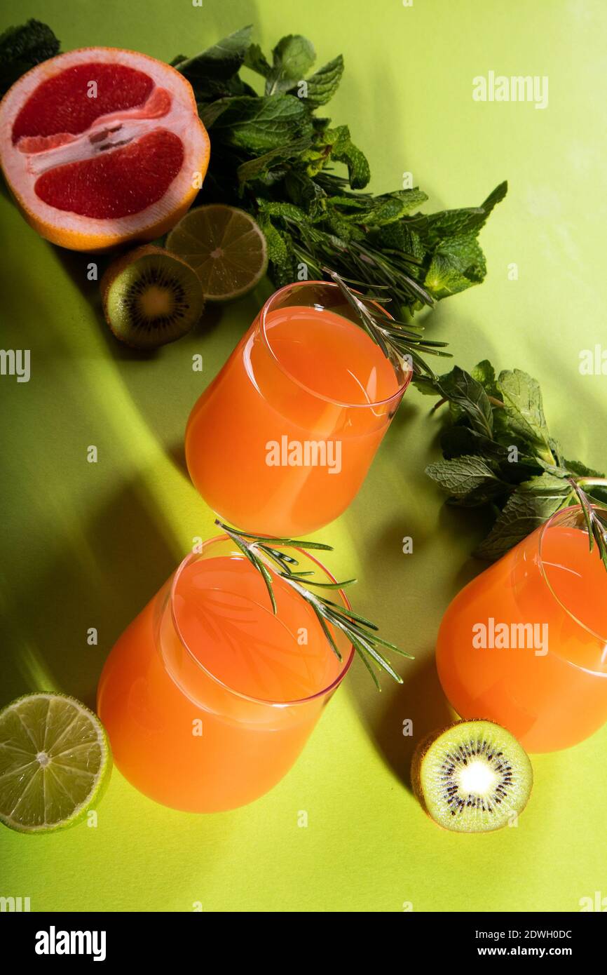Composizione di bicchieri con succo d'arancia, foglie di arancio, lime, kiwi, rosmarino e menta tagliate su sfondo verde chiaro Foto Stock