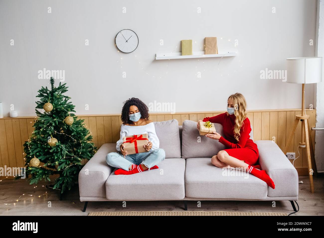 Festeggiamo Natale e Capodanno 2021 durante il concetto pandemico di Covid-19. Due donne multietniche in maschere protettive sedute sul divano e scambiate Foto Stock