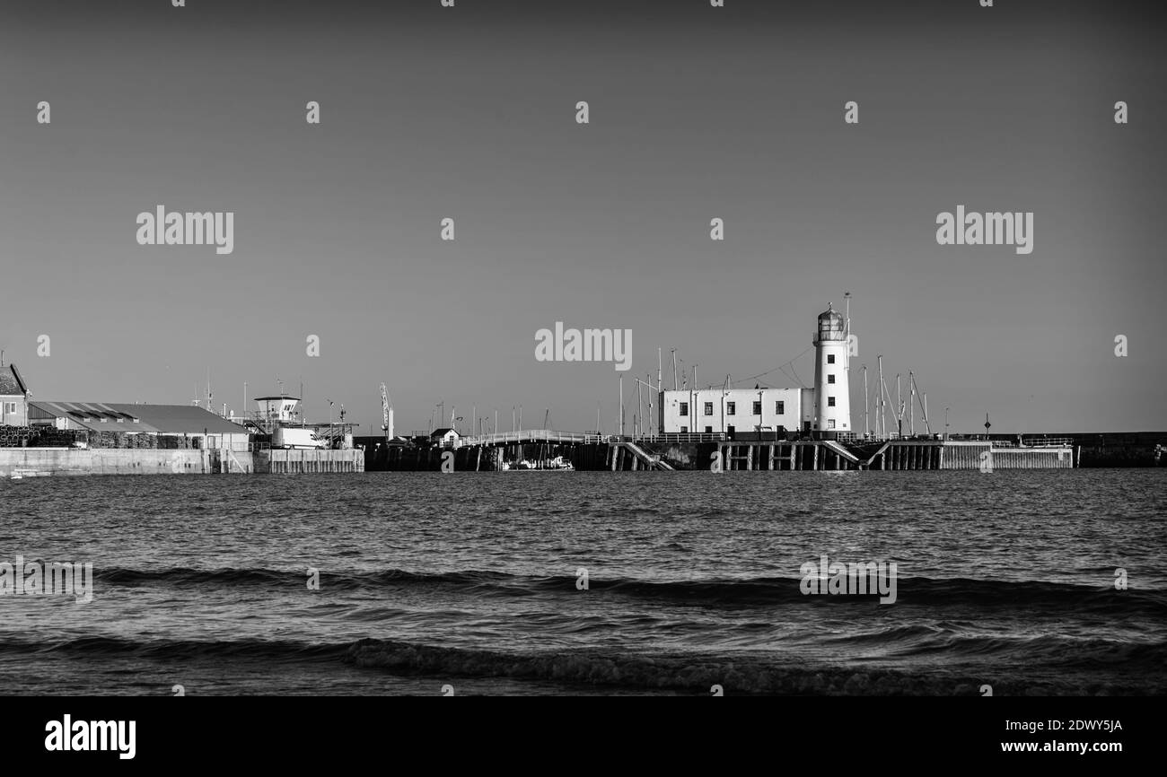Le onde si rotolano delicatamente verso una spiaggia e un faro si erge su un molo che si estende verso il mare. Foto Stock